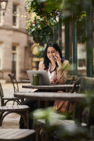 Mulher jovem profissional sorrindo enquanto fala ao telefone em café ao ar livre, com laptop aberto em mesa de madeira, bebida verde à frente, plantas trepadeiras ao fundo e fachada de prédio urbano desfocada