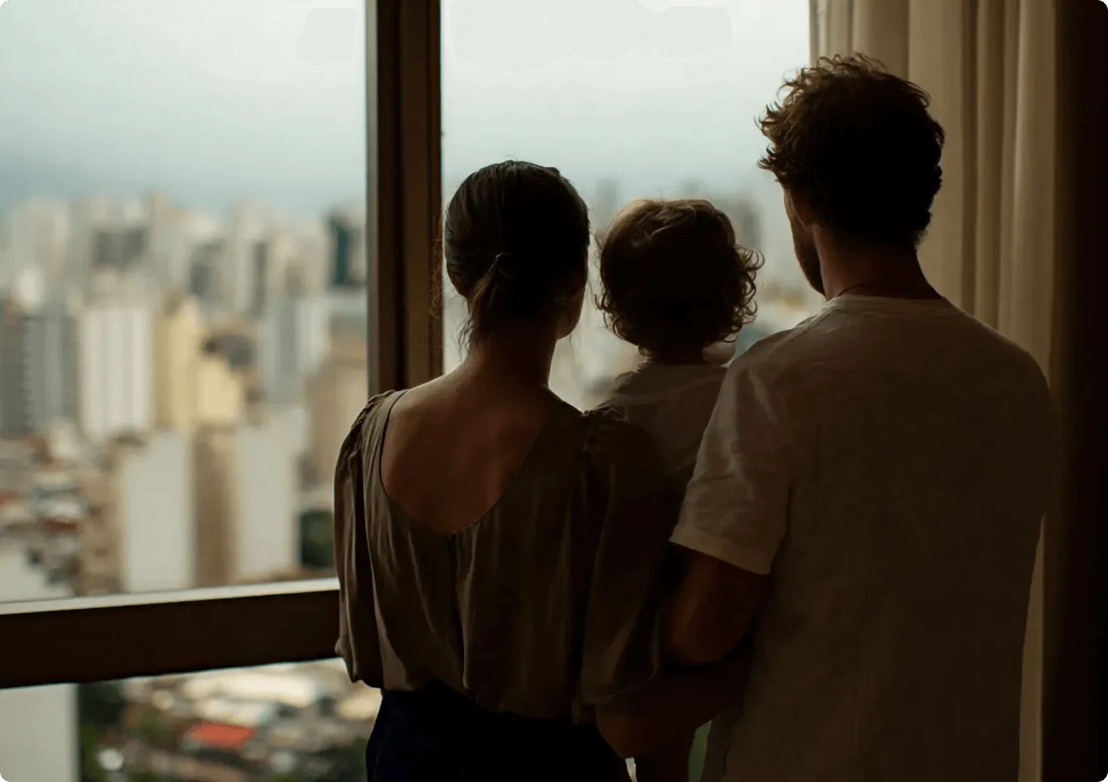 Homem adulto, mulher adulta e criança vistos de costas observando paisagem urbana através de grande janela em apartamento de prédio alto, com vista de edifícios e vista cityscape ao fundo desfocada