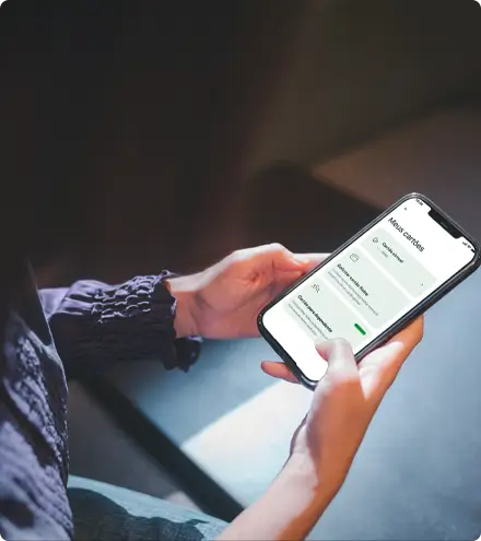 Pessoa segurando smartphone exibindo interface do app de cartão Avenue com informações visíveis de transações ou saldo, vistindo roupas em tons azuis e cinzentos em ambiente externo com iluminação azulada suave, transmitindo acesso móvel fácil a serviços bancários e de cartão