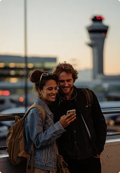 Casal jovem sorridente em terminal de aeroporto consultando smartphone juntos, vistindo jaquetas de lã em tons pretos e cinzentos com mochilas ao ombro, com vista para torre de controle do aeroporto e céu em tons dourados e rosados ao pôr do sol, transmitindo viagem internacional e acesso a serviços de viagem