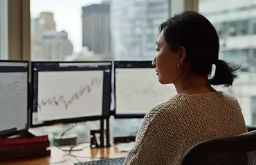 Profissional analisa gráficos financeiros em dois monitores em escritório com vista para a cidade.
