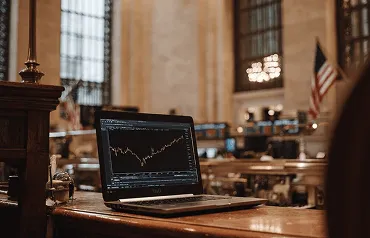Notebook com gráfico financeiro sobre mesa em amplo salão histórico, com bandeira americana ao fundo.