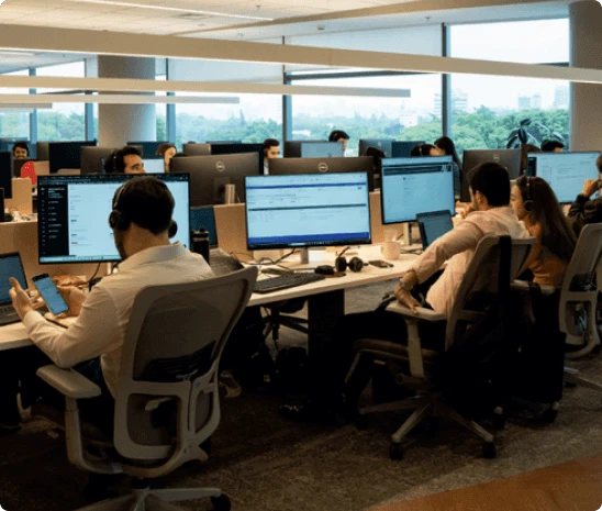 Centro de atendimento ou customer service mostrando múltiplos profissionais em estações de trabalho individuais equipadas com dois monitores cada, em escritório corporativo moderno com grandes janelas panorâmicas oferecendo vista urbana e paisagem, pé direito alto com estrutura de vigas em tons neutros, iluminação natural clara, transmitindo equipe dedicada e profissional de suporte ao cliente