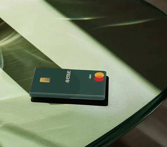 Cartão de débito internacional em verde escuro da marca Avenue exibindo logo "avenue" em branco, chip de segurança EMV em tom dourado no canto superior esquerdo, bandeira de pagamento Mastercard em laranja e vermelho no canto inferior direito, design minimalista sobre superfície em tons verdes com iluminação lateral dramática criando contraste visual