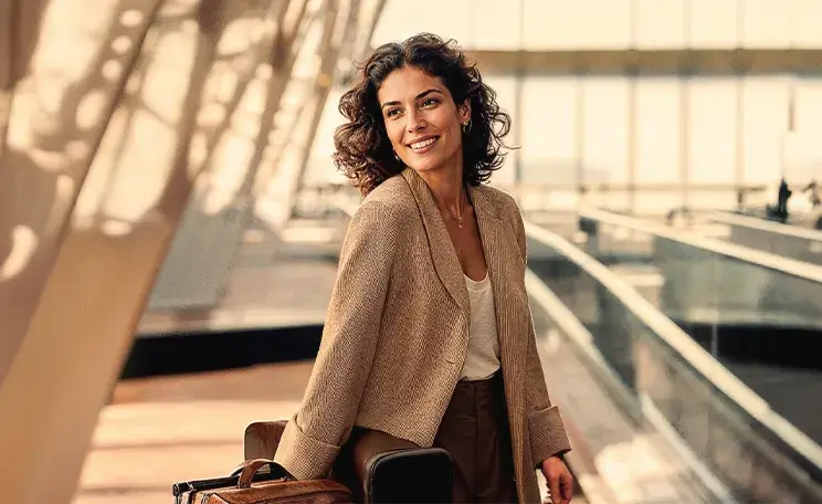 Mulher profissional com cabelo cacheado e mala de viagem, sorrindo em ambiente moderno com grandes janelas de vidro, luz natural intensa, sugerindo aeroporto ou terminal de viagem internacional