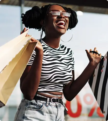 Mulher jovem sorrindo alegremente enquanto carrega várias sacolas de compras em cores vibrantes, usando óculos e camiseta preta e branca com padrão de zebra, em ambiente de rua com luz natural clara