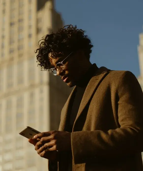 Homem jovem com cabelo cacheado usando óculos de sol e jaqueta bege, segurando smartphone enquanto observa, em ambiente urbano com prédios ao fundo e céu azul claro