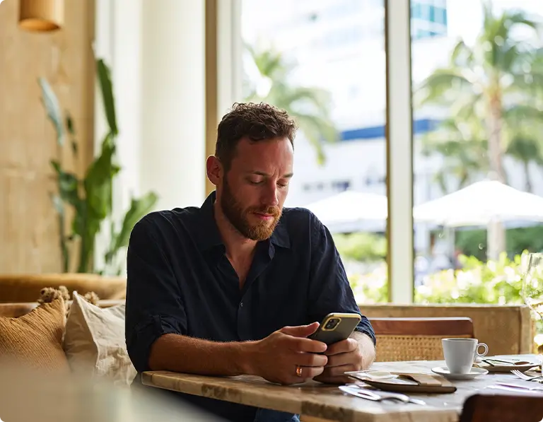 Homem profissional com barba consultando smartphone em ambiente de resort tropical sofisticado, vistindo camisa preta, sentado à mesa de madeira com xícara de café branca e documentos, grande janela mostrando vista para praia, palmeiras, guarda-sol branco e céu azul claro, transmitindo flexibilidade de trabalho em renda fixa de alto padrão