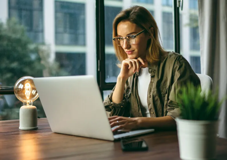 Mulher trabalhando concentrada em um laptop, em mesa de escritório com iluminação aconchegante.