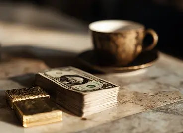 Natureza morta artística com xícara de café desfocada ao fundo, maço de notas de dólar americano em primeiro plano e pequena barra de ouro em superfície cinza, iluminação lateral dramática com sombras acentuadas
