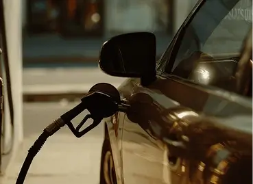 Close-up de bico de bomba de combustível preto inserido em tanque de carro marrom durante abastecimento, com iluminação de posto de gasolina ao fundo desfocada