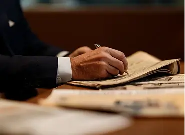 Mão de profissional em paletó preto sobre documento ou contrato em mesa de madeira durante assinatura ou análise, com copo preto e documentos desfocados ao fundo